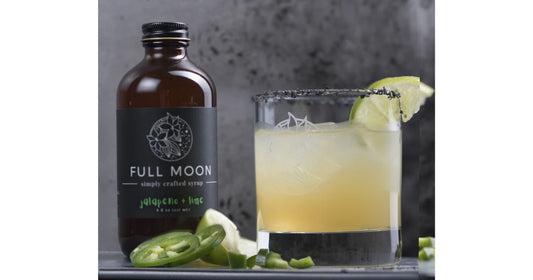 Spicy jalapeño margarita with black salt rim and lime garnish next to a bottle of Full Moon jalapeño lime simple syrup