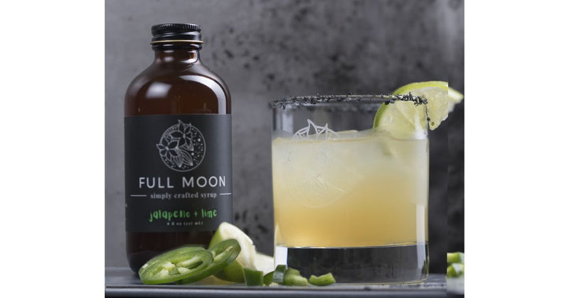 Spicy jalapeño margarita with black salt rim and lime garnish next to a bottle of Full Moon jalapeño lime simple syrup